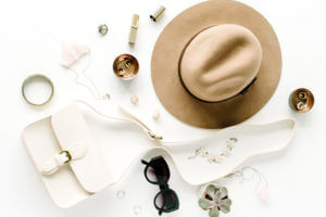 Brown hat, white purse, jewelry, and black sunglasses laid out on table