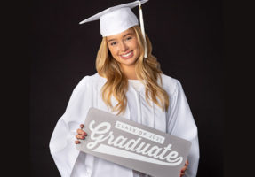 Girl smiling wearing white graduation cap and gown holding sign that says graduate