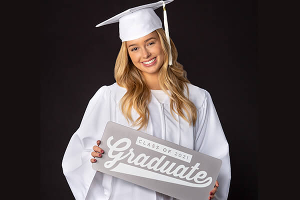 Girl smiling wearing white graduation cap and gown holding sign that says graduate