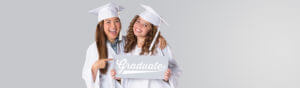 Girls smiling wearing white graduation cap and gown holding sign that says graduate